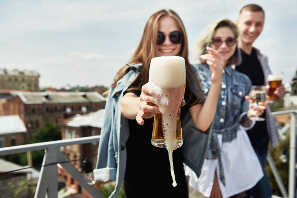 Close up of glass of beer with froth in girl hand with group of friends in blur on background - Photo, Image