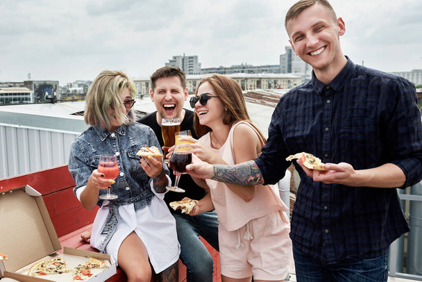 Group of friends drinking beer and eating pizza before festival at outdoors pub on roof, toasting and laughing - Foto, imagen
