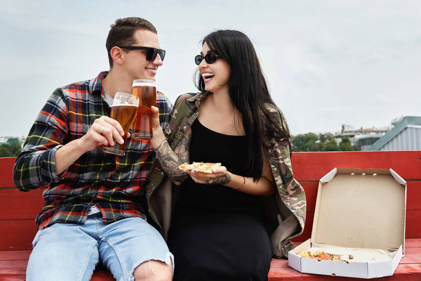 Happy smiling couple eating pizza, toasting and drinking beer at outdoor pub or bar on roof - 写真・画像