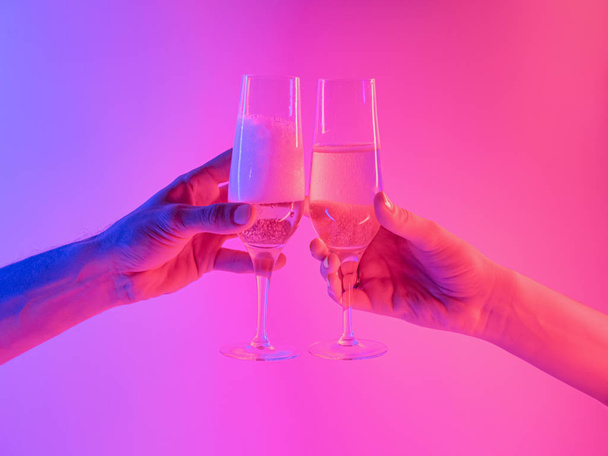 Partying, celebration concept: two glasses of sparkling wine in hands in bright neon background.  - Photo, image