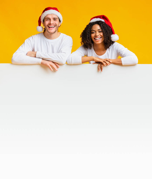サンタの異人種間のカップルは白い空の広告ボードでリース帽子. - 写真・画像