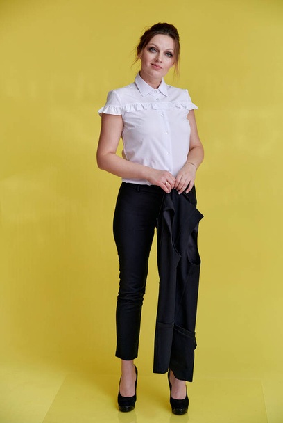 Business concept. Full-length portrait of an adult pretty woman of 40 years old with good makeup in a business suit on a yellow background. Standing right in front of the camera with a smile. - Fotoğraf, Görsel
