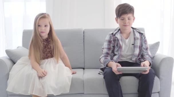 Portrait of two Caucasian children sitting on couch. Boy watching something at tablet screen, girl in beautiful dress looking away. First love, leisure. - Imágenes, Vídeo