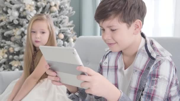 Side view of happy Caucasian boy sitting with tablet and smiling, pretty girl looking at him at the background. Shy cute child sitting next to her love on New Years eve. First love, childhood. - Filmati, video