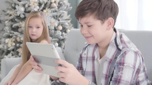 Portrait of cute Caucasian girl sitting at the background and looking at boy playing with tablet. Shy little lady in beautiful dress and brunette boy resting on couch on Christmas eve. Childhood. - Filmati, video