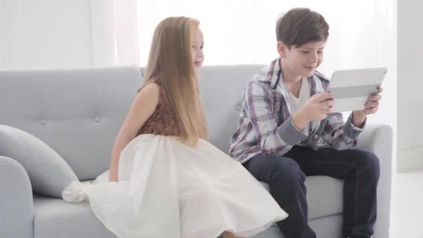 Little cute Caucasian girl moving closer to boy watching movie on tablet screen. Shy girl in dress waiting for attention from her love. Childhood, first love, leisure. - Кадри, відео