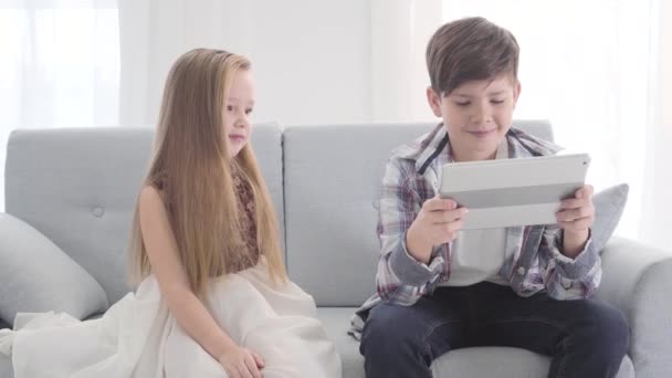 Charming Caucasian girl sitting on couch with boy holding tablet. Shy pretty little lady looking at her love. Camera slowly moving around people from left to right. - Video