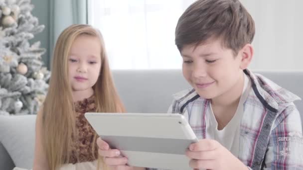 Close-up face of smiling Caucasian boy holding tablet as pretty girl looking at him from the background. Children resting on couch in front of Christmas tree. Leisure, first love, happiness. - Filmagem, Vídeo