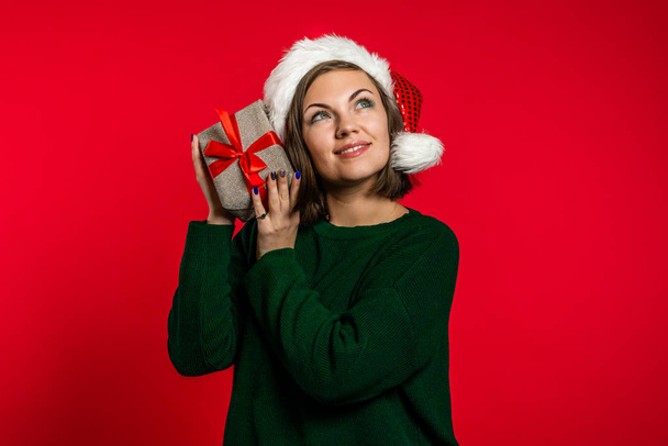 Молода жінка посміхається і тримає подарункову коробку біля вуха, щоб вгадати, що всередині на червоному студійному фоні. Дівчина в капелюсі Санта. Різдвяний настрій
. - Фото, зображення