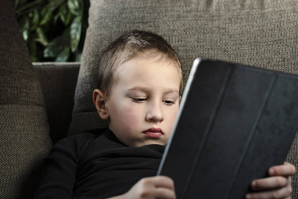 Молодий хлопчик сидить на дивані у вітальні і дивиться мультфільми на планшеті
 - Фото, зображення