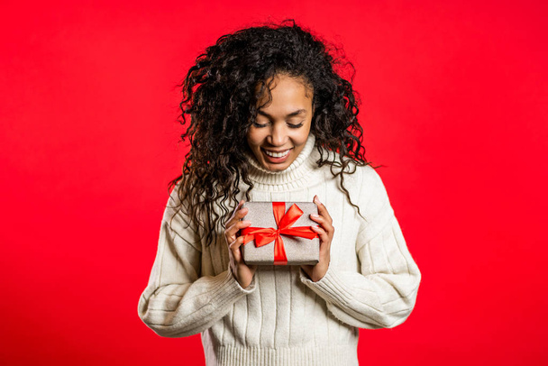 Young pretty african woman smiling and holding gift box on red studio background. Girl with curly hairstyle in white sweater. Christmas mood. - 写真・画像