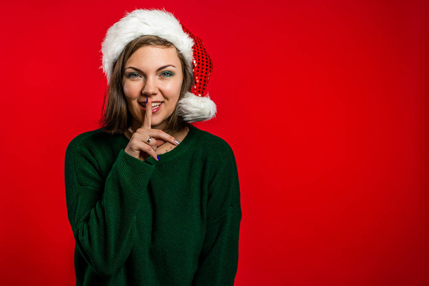 Beautiful woman in Christmas Santa hat holding finger on her lips over red background. Gesture of shhh, secret, silence. Close up. - Fotografie, Obrázek