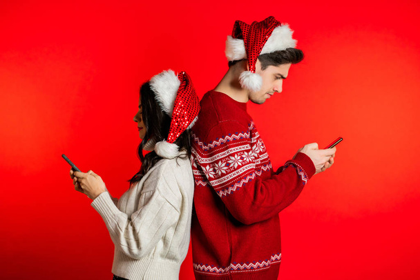 Young couple in Santa hats stand with their backs to each other and print messages or surf the Internet from their mobile phone. Technology, smartphone, social networks concept - Foto, afbeelding