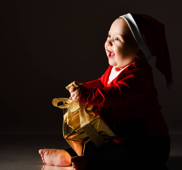 Infant boy toddler in red santa claus costume and barefooted is holding trying to untie new year gift box present - Zdjęcie, obraz