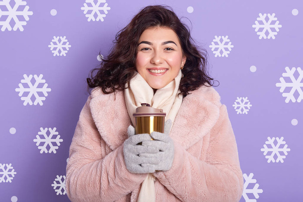 Close up portrait of adorable smiling woman wearing pale pink faux fur coat and, holding thermo mug with coffee or tea, drinking hot beverage, posing isolated over wall with snowflakes on background. - Photo, Image