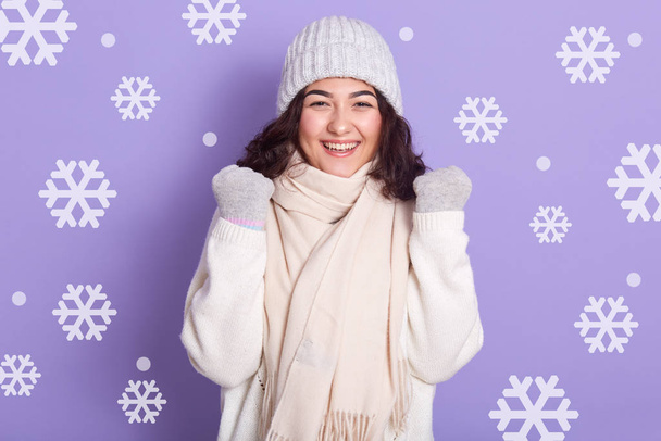 Image of beautiful girl stands smiling and clenches her fists, celebrating sucsses, wearing winter clothes, looks at camera, has dark hair, studio photo isolated over lilac background with snowflakes. - Photo, Image
