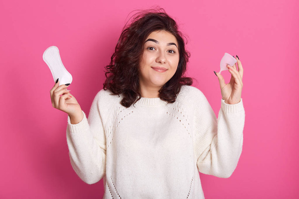 Closeup portrait of confused surprised female with curly hair holding hygiene pad and menstrual cup, having periods, making choice, standing isolated over pink background in studio. Hygiene concept. - Фото, изображение