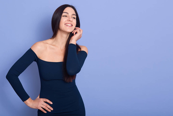Smiling elegance woman wearing dark blue dress standing and looking away. Beuitiful brunette female keeps one hand on hip and another near face, smiling girl, copy space foe adverisment or promotion. - Zdjęcie, obraz