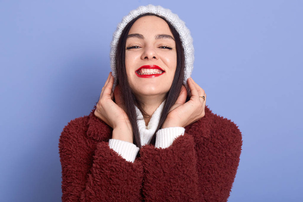 Picture of magnetic emotional brunette with red lips wearing white hat and fauxfur coat, having pleasant facial expression, smiling sincerely, being in high spirits. People and emotions concept. - Photo, image