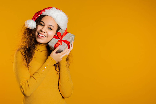 Eine aufgeregte Frau mit Weihnachtsmann-Hut erhielt eine Geschenkbox mit Schleife. Sie ist glücklich und von der Aufmerksamkeit geschmeichelt. Mädchen mit Geschenk auf gelbem Hintergrund. Studioaufnahme - Foto, Bild