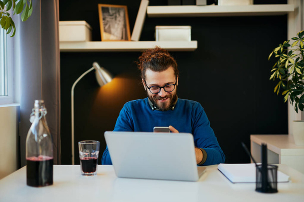 Atraente sorrindo positivo barbudo freelancer sentado em seu escritório em casa e usando telefone inteligente para ler ou enviar mensagem
. - Foto, Imagem