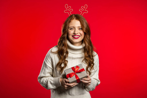 Young pretty woman smiling and holding gift box on red studio background. Girl with beautiful hairstyle in white sweater and deer horns. Christmas mood. - Fotoğraf, Görsel