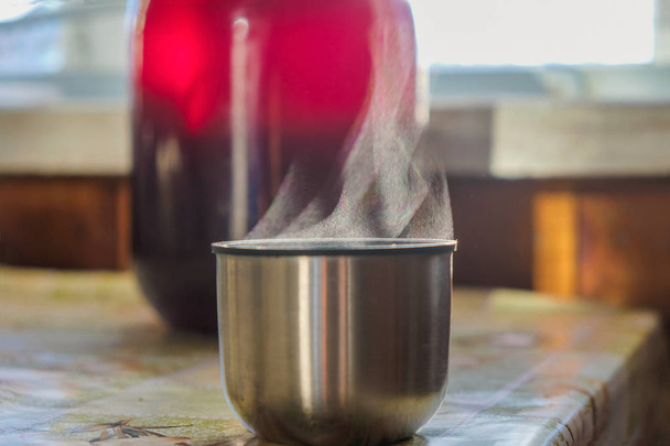 Párolgás. Meleg gőzölgő termosz az asztalon egy hideg napon Ukrajnában. Vidéki csendélet egy üveg házi borral és egy csészével.. - Fotó, kép