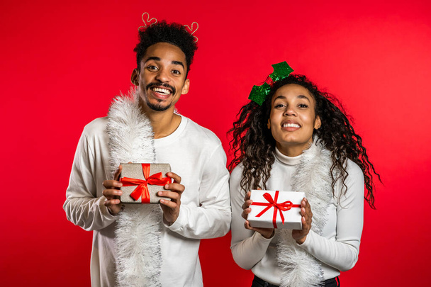 Young african american couple with christmas gift box isolated on red background studio. New 2020 year decorations, presents, party, happiness concept. - Photo, image