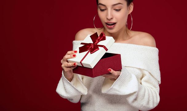 The girl eagerly opens the gift box on a red background. - Photo, Image