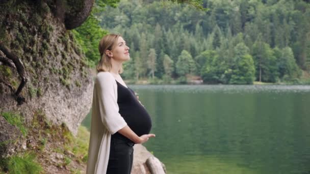 Беременная женщина нежно касается живота, стоя у горного озера.
. - Кадры, видео