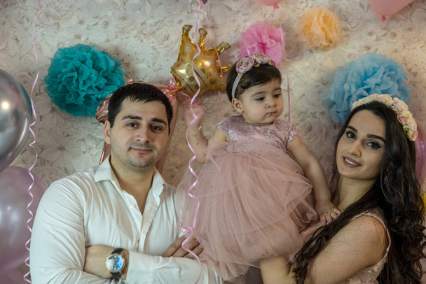 Baby's birthday celebration. The girl was one year old. Happy parents. The child looks away. All are dressed in elegant clothes. Young, beautiful married couple and a child of Caucasian ethnicity. Against the background of balloons, in pink colors. - Valokuva, kuva
