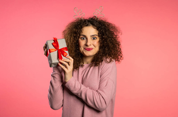 Cheerful excited woman with curly hair smiling and holding gift box on pink studio background. Cute girls portrait in deer horns wreath. Christmas mood. - Фото, изображение