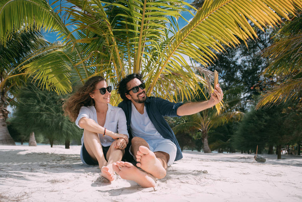 A pair of lovers take a selfie under a palm tree on a tropical beach - Φωτογραφία, εικόνα
