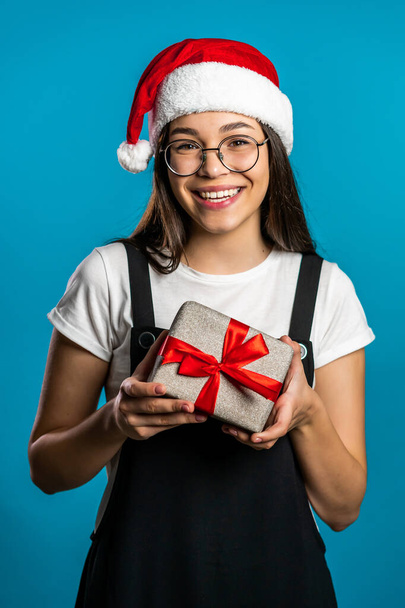 Pretty young girl smiling and holding gift box on blue studio background. Cute portrait in Santa hat. Christmas mood. - Фото, зображення