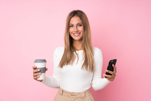 Jeune femme blonde sur fond rose isolé tenant du café à emporter et un mobile
 - Photo, image