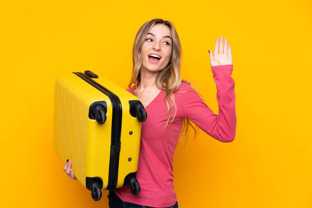 Young woman over isolated yellow background in vacation with travel suitcase and saluting - Foto, afbeelding