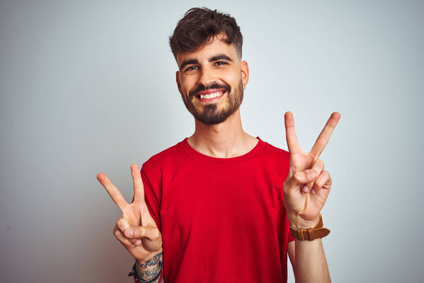 Jongeman met tatoeage in rood t-shirt staat over een geïsoleerde witte achtergrond glimlachend naar de camera te kijken met vingers die een overwinningsteken doen. Nummer twee. - Foto, afbeelding