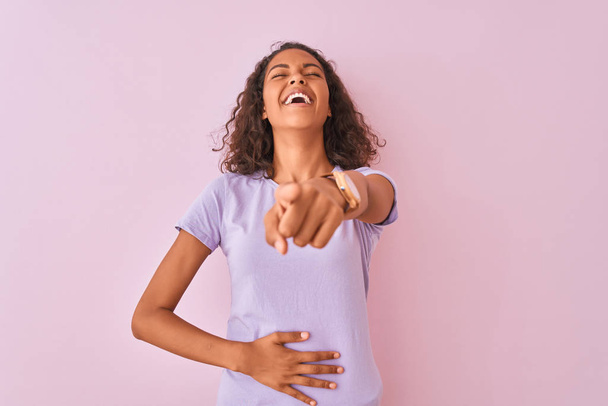 Mujer brasileña joven con camiseta de pie sobre un fondo rosa aislado riéndose de ti, señalando con el dedo a la cámara con la mano sobre el cuerpo, expresión de vergüenza
 - Foto, Imagen