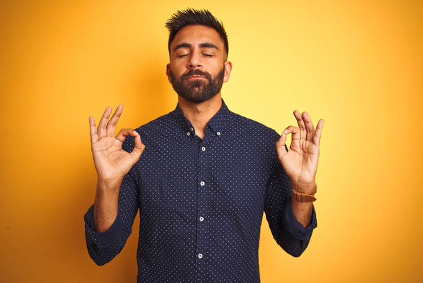 Fiatal jóképű indiai üzletember inget visel elszigetelt sárga háttér pihenni és mosolyogni csukott szemmel csinál meditációs gesztus ujjakkal. A jóga fogalma. - Fotó, kép