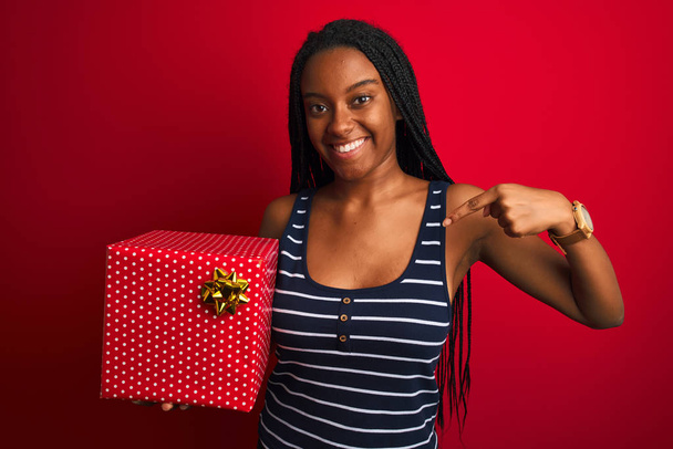 jong Afrikaans amerikaans vrouw met verjaardag geschenk staande over geïsoleerde rode achtergrond met verrassing gezicht wijzend vinger naar zichzelf - Foto, afbeelding