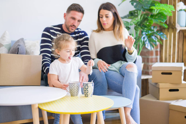 Mooie familie, ouders zittend op de Bank drinken koffie op zoek zijn kind spelen in nieuw huis rond kartonnen dozen - Foto, afbeelding