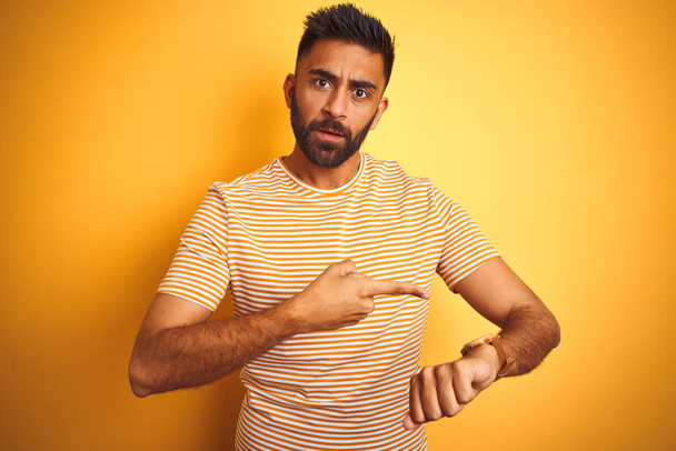 Jeune homme indien portant t-shirt debout sur fond jaune isolé Dans la hâte pointant vers regarder le temps, l'impatience, bouleversé et en colère pour retard de délai
 - Photo, image