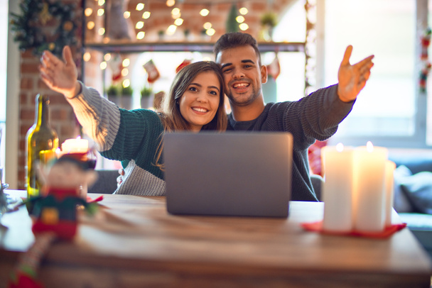 Nuori kaunis pari istuu kannettavan tietokoneen ympärillä joulukoristeita kotona katsoen kameraa hymyillen avosylin halausta varten. Iloinen ilme, johon kuuluu onnellisuus
. - Valokuva, kuva