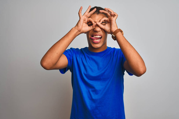 Jeune homme arabe beau portant un t-shirt bleu debout sur fond blanc isolé faisant un geste correct comme des jumelles sortant la langue, les yeux regardant à travers les doigts. Expression folle
. - Photo, image