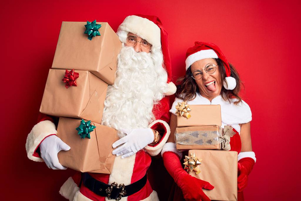 Middle age couple wearing Santa costume holding tower of gifts over isolated red background sticking tongue out happy with funny expression. Emotion concept. - Photo, Image