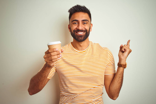 junger indischer Mann, der eine Tasse Kaffee trinkt, steht vor isoliertem weißen Hintergrund und zeigt sehr glücklich mit der Hand und dem Finger zur Seite - Foto, Bild