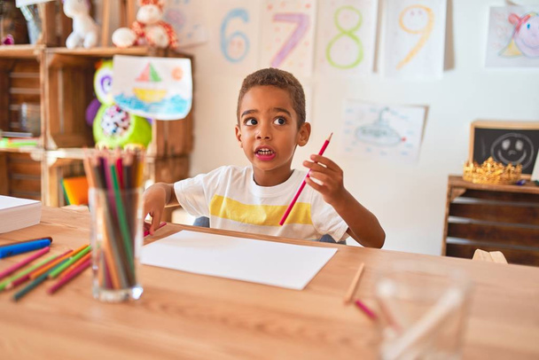 Hermoso niño afroamericano sentado dibujo usando papel y lápices en el escritorio en el jardín de infantes
 - Foto, Imagen