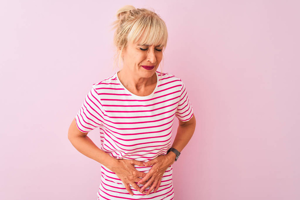 Frau mittleren Alters in gestreiftem T-Shirt vor isoliertem rosa Hintergrund mit der Hand auf dem Bauch wegen Verdauungsstörungen, schmerzhafte Krankheit, die sich unwohl anfühlt. Schmerzkonzept. - Foto, Bild