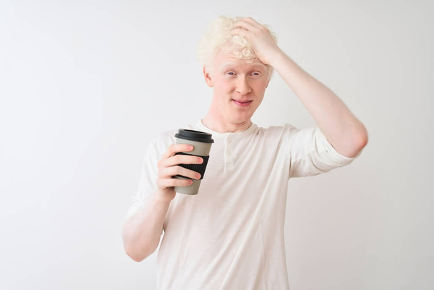 Mladý blonďatý albín, který pije kávu, stojí nad osamělým bílým pozadím, vystresovaný rukou na hlavě, šokovaný hanbou a překvapenou tváří, rozzlobený a frustrovaný. Strach a zmatek z omylu. - Fotografie, Obrázek