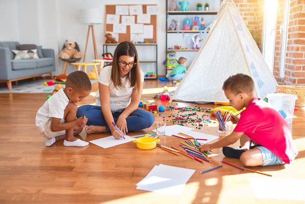 Jovem e bela professora e crianças sentadas no chão desenhando em torno de muitos brinquedos no jardim de infância
 - Foto, Imagem